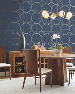 A modern dining room featuring a wooden table and chairs, York Wallcoverings’ Sun Circles Wallpaper in blue with geometric patterns, a gold chandelier, a white orchid centerpiece, and stylish decor on the sideboard.