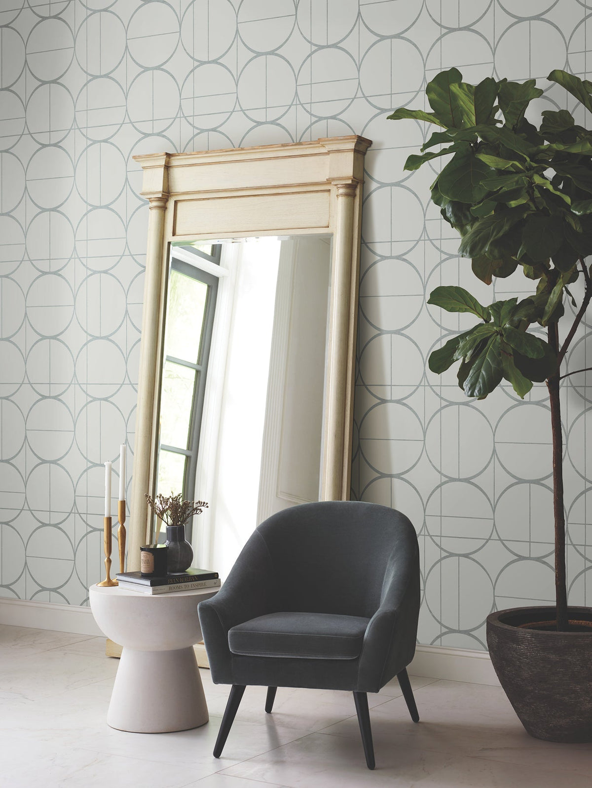 A gray armchair sits near a small white table with decor, a large ornate mirror, and a tall potted plant against a wall adorned with York Wallcoverings’ Sun Circles Wallpaper. Natural light comes through a window reflected in the mirror.
