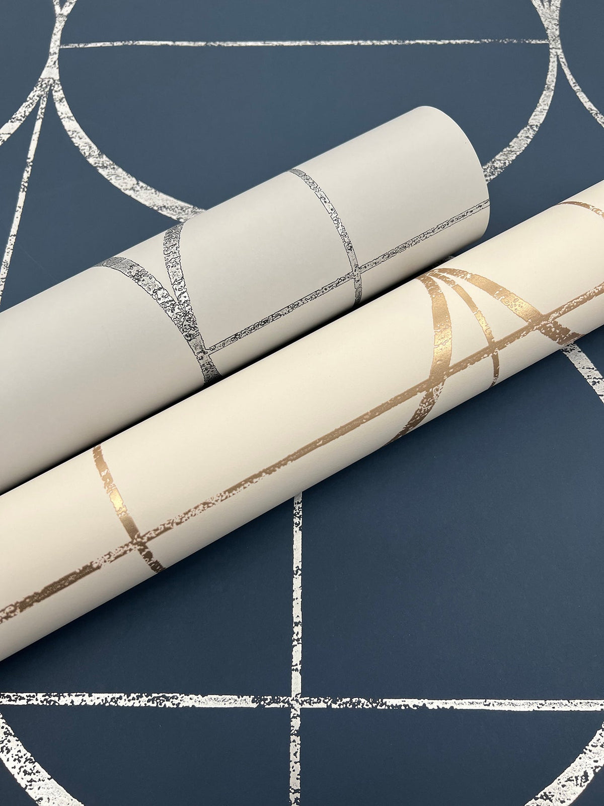 Two rolls of York Wallcoverings’ Sun Circles Wallpaper—one in silver and one in gold, both featuring metallic geometric patterns—are displayed on a dark blue background with a matching metallic geometric design.