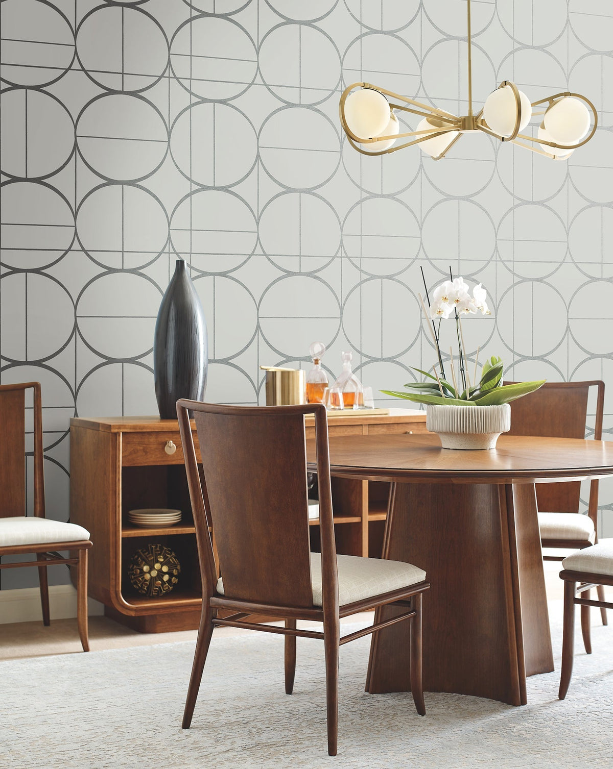 Mid-century modern dining room featuring a wood table, four chairs, a sideboard, York Wallcoverings’ Sun Circles Wallpaper, a gold chandelier, a tall black vase, and a white orchid centerpiece on the table.