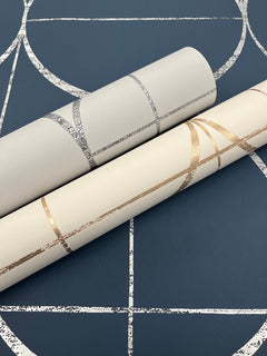 Two rolls of Sun Circles Wallpaper by York Wallcoverings, featuring metallic geometric patterns, rest on a dark blue surface adorned with a similar silver geometric design.