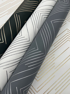 Three rolls of York Wallcoverings’ Polished Chevron Wallpaper, in black, white, and gray colorways, each featuring metallic chevron patterns in gold or silver, arranged diagonally on a flat surface.
