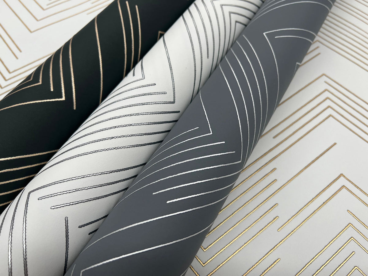 Three rolls of York Wallcoverings’ Polished Chevron Wallpaper featuring geometric metallic lines in gold and silver on black, white, and gray backgrounds are arranged diagonally atop a matching patterned surface.