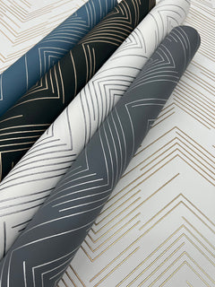 Four rolls of Polished Chevron Wallpaper by York Wallcoverings in blue, black, white, and gray tones with metallic lines are arranged diagonally on a sheet featuring a similar gold and white chevron pattern.