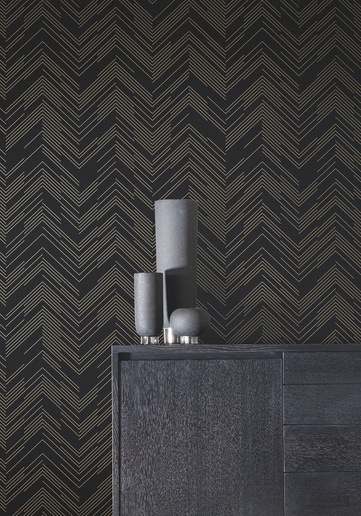 A minimalist black sideboard is styled with three matte black decorative vases of varying heights, set against York Wallcoverings’ Polished Chevron Wallpaper in black with a striking gold chevron line pattern.