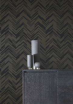 A minimalist black sideboard is styled with three matte black decorative vases of varying heights, set against York Wallcoverings’ Polished Chevron Wallpaper in black with a striking gold chevron line pattern.