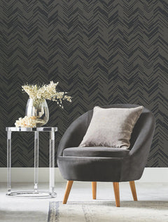 A modern gray chair with a cushion sits next to a clear round side table holding a glass vase with white flowers, set against York Wallcoverings’ Polished Chevron Wallpaper in black.