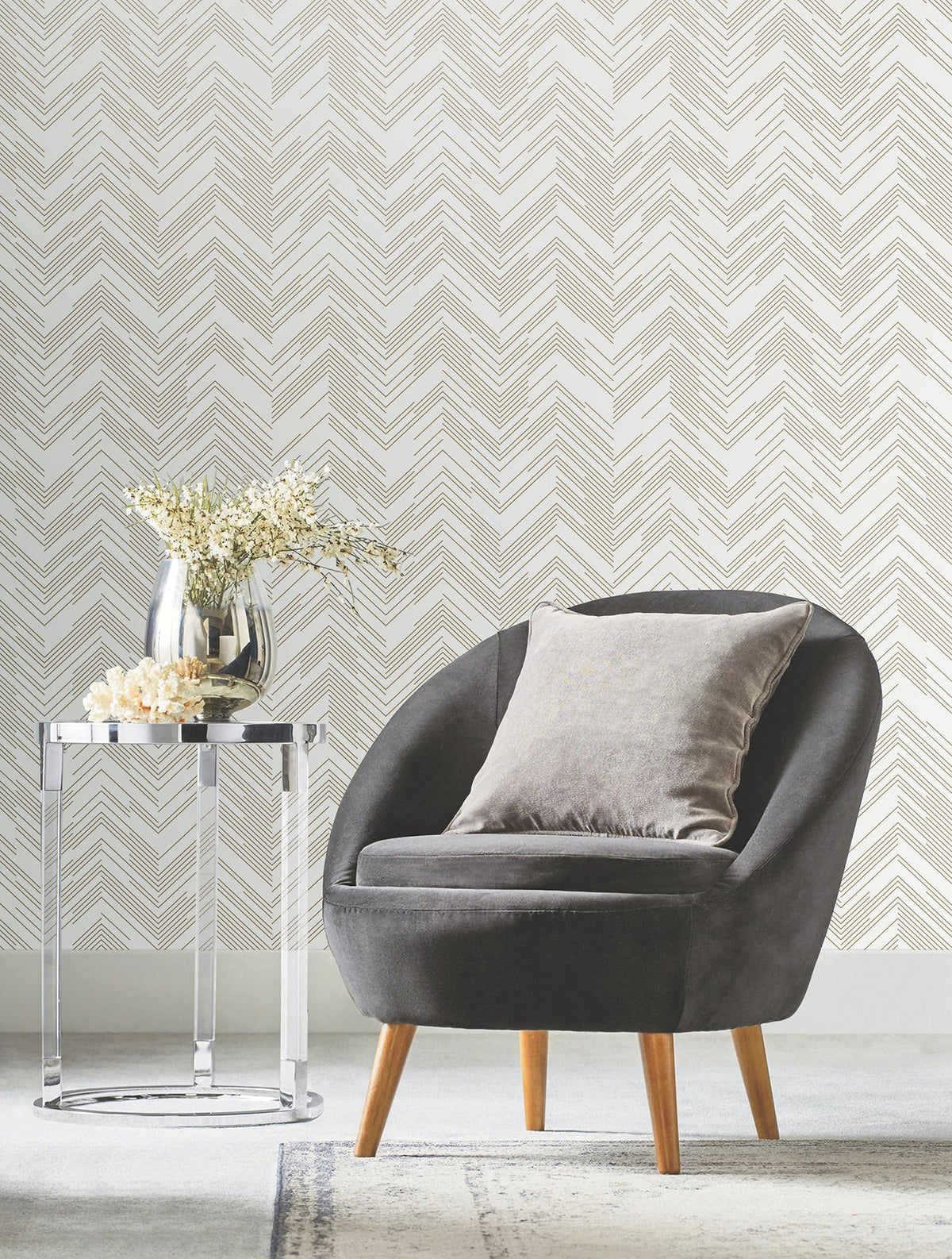 A gray armchair with a pillow sits beside a round glass side table holding a vase of flowers, set against a white wall adorned with York Wallcoverings’ Polished Chevron Wallpaper.