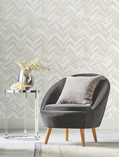 A gray armchair with a pillow sits beside a round glass side table holding a vase of flowers, set against a white wall adorned with York Wallcoverings’ Polished Chevron Wallpaper.