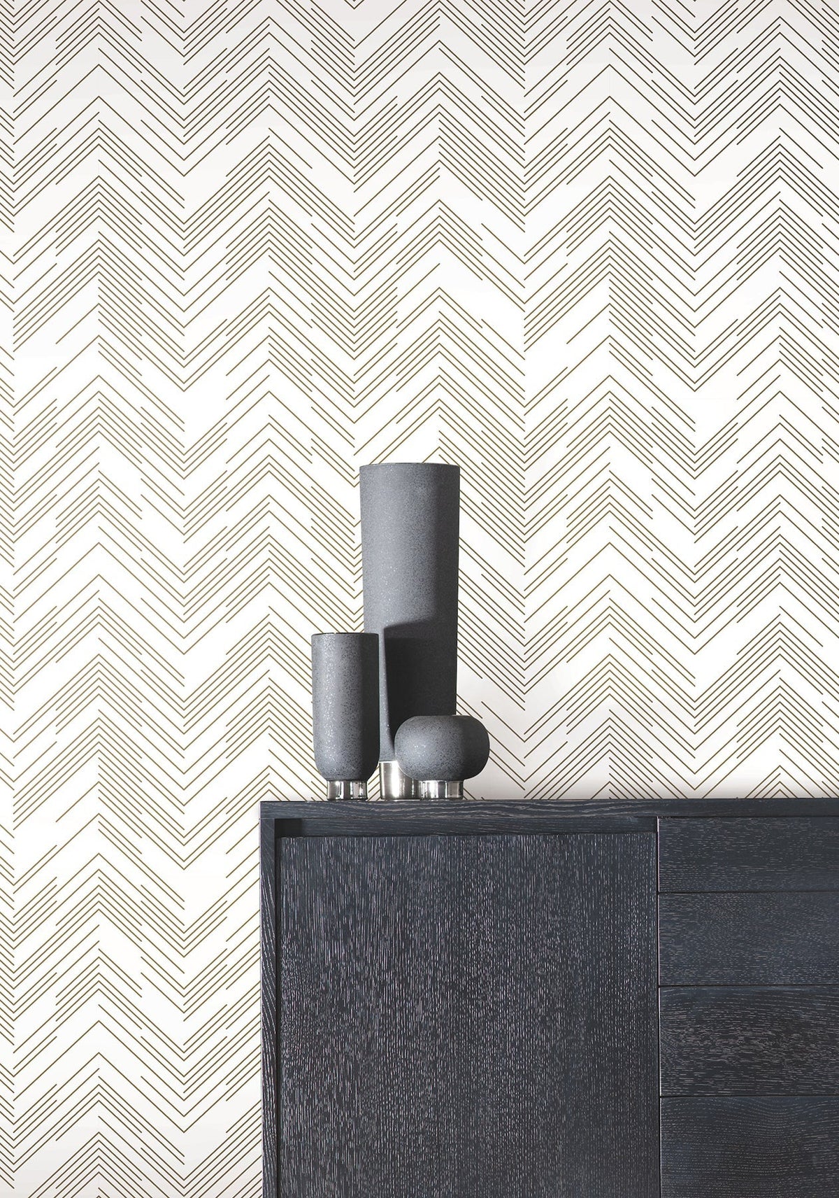 A black cabinet with three matte vases sits in front of a white wall adorned with the Polished Chevron Wallpaper by York Wallcoverings. The scene is modern and minimal.