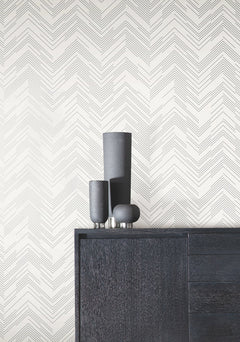 A dark wooden cabinet with three black decorative vases sits in front of a white wall adorned with York Wallcoverings’ Polished Chevron Wallpaper.