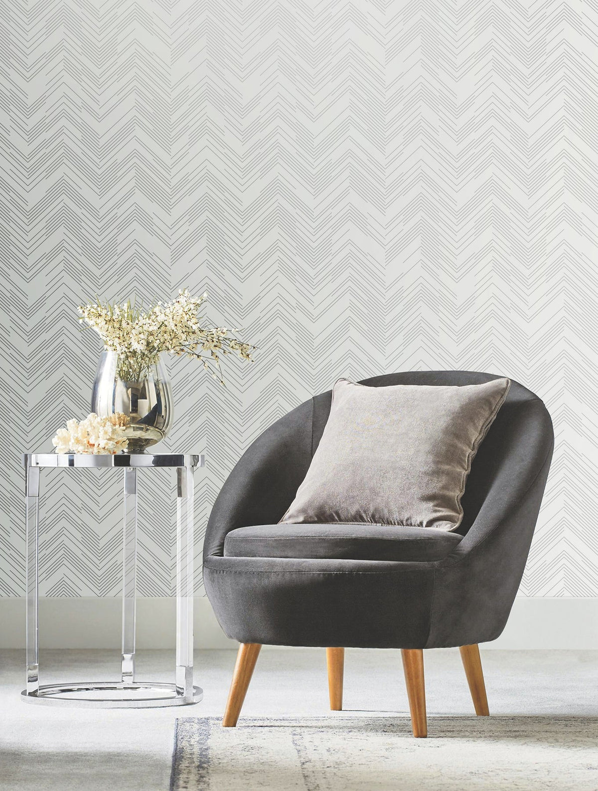 A modern gray accent chair with a pillow sits next to a round metal side table holding a vase of white flowers, set against York Wallcoverings’ Polished Chevron Wallpaper and a light rug.