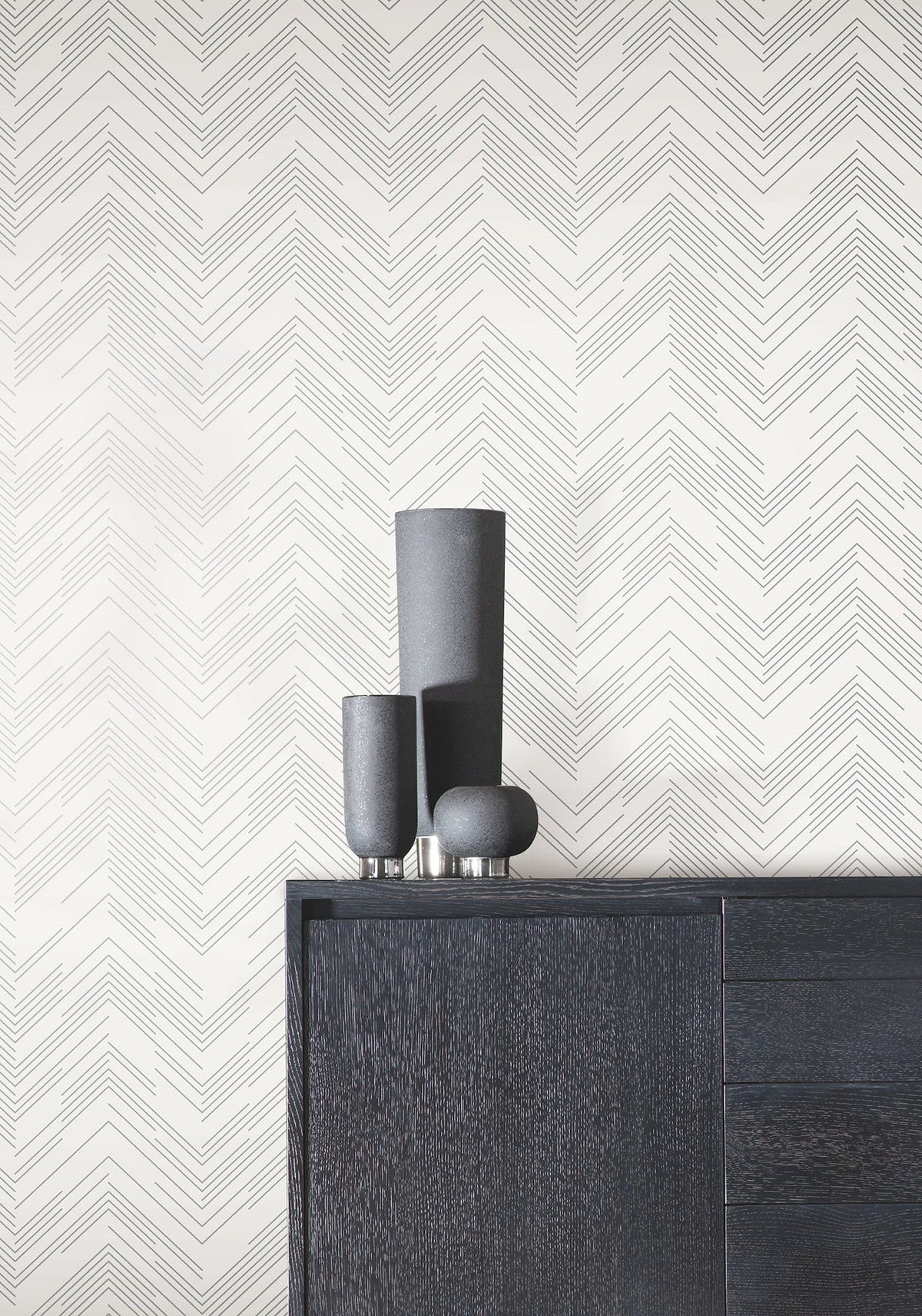 A dark wooden cabinet with three matte black vases sits in front of a white wall adorned with York Wallcoverings’ Polished Chevron Wallpaper.