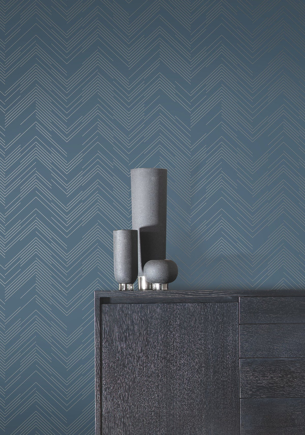 Three modern gray vases sit on a dark wooden cabinet against a blue wall adorned with York Wallcoverings’ Polished Chevron Wallpaper. The scene is minimalist and contemporary.