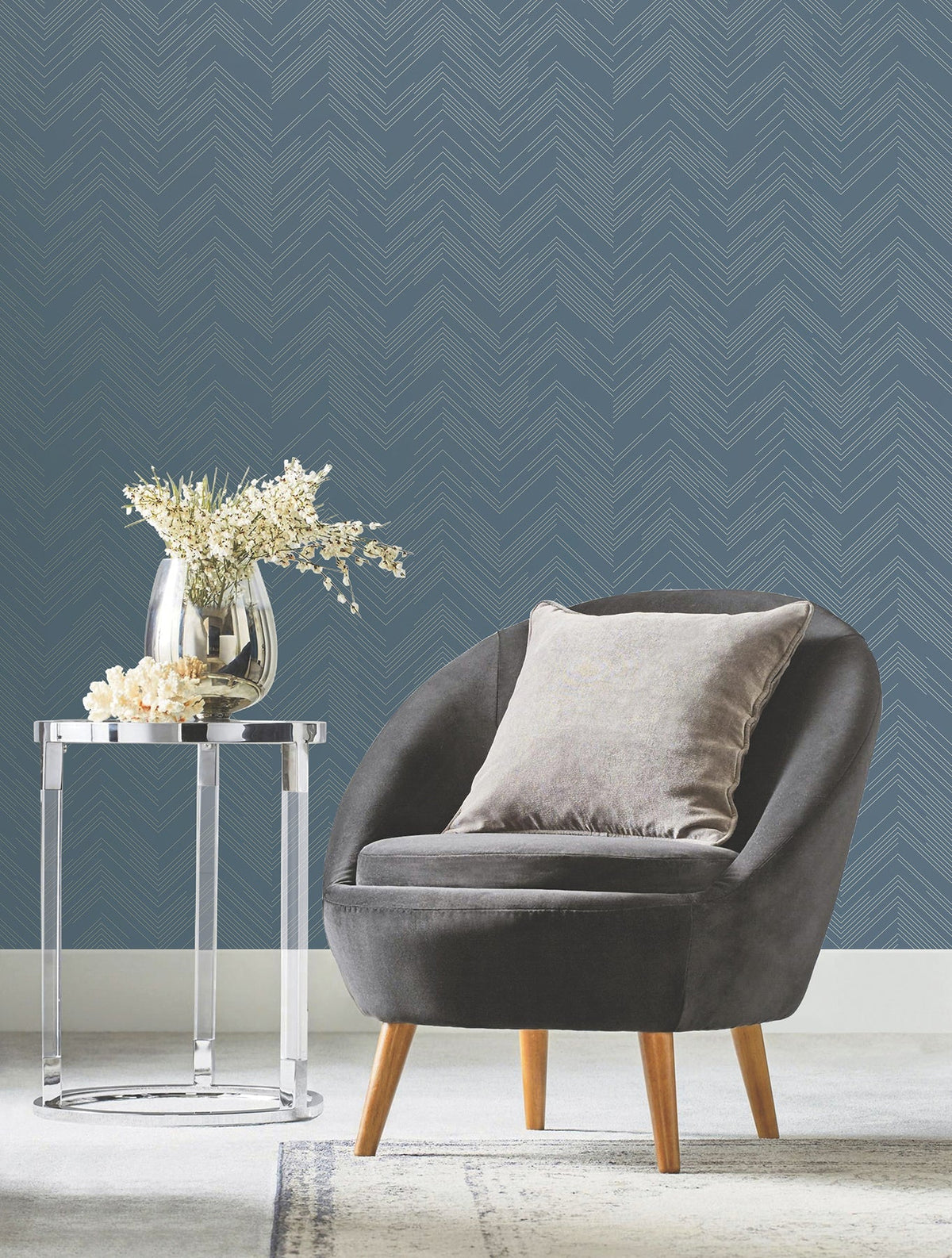 A modern gray chair with a plush pillow sits next to a clear side table holding a vase with white flowers, set against York Wallcoverings’ Polished Chevron Wallpaper and a light carpet.