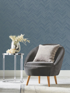 A modern gray chair with a plush pillow sits next to a clear side table holding a vase with white flowers, set against York Wallcoverings’ Polished Chevron Wallpaper and a light carpet.