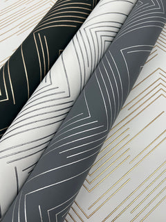 Three rolls of the York Wallcoverings Polished Chevron Wallpaper in black, white, and gray, each featuring metallic geometric chevron patterns in gold or silver, are arranged diagonally on a matching patterned surface.