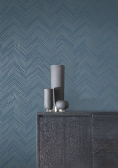 Three matte black vases of varying heights sit on a dark wooden cabinet against a blue wall adorned with Polished Chevron Wallpaper by York Wallcoverings.