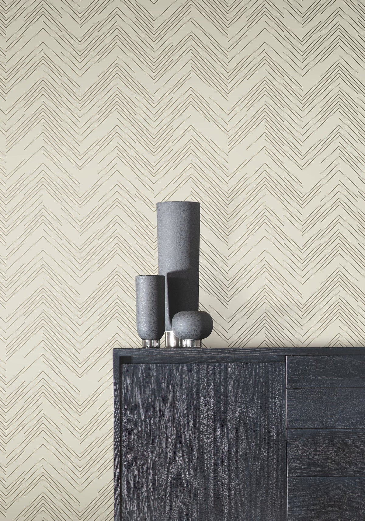 A modern black cabinet holds three minimalist black vases in front of a beige wall adorned with York Wallcoverings’ Polished Chevron Wallpaper.