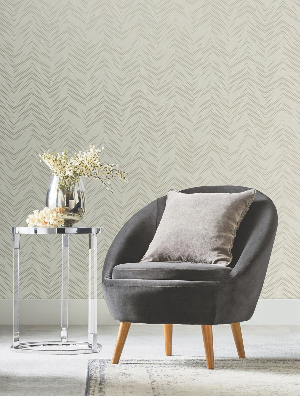 A modern gray chair with a gray pillow sits beside a round chrome side table holding a silver vase with white flowers, set against York Wallcoverings’ Polished Chevron Wallpaper.