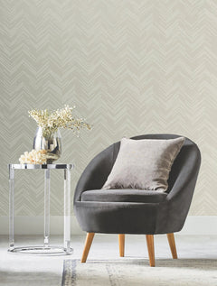 A modern gray chair with a gray pillow sits beside a round chrome side table holding a silver vase with white flowers, set against York Wallcoverings’ Polished Chevron Wallpaper.