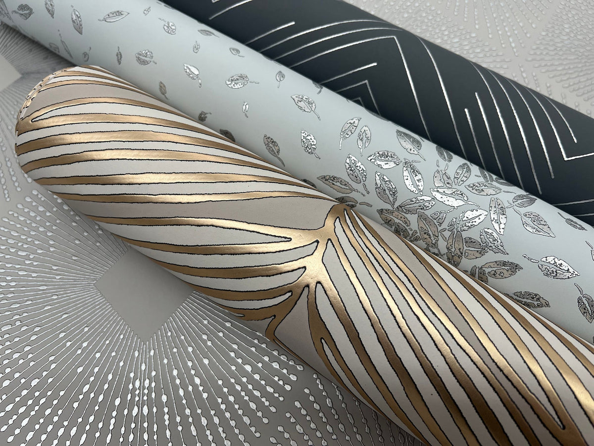 Three rolls of York Wallcoverings Polished Chevron Wallpaper, featuring metallic designs: one with gold and white abstract lines, one adorned with silver leaves on light gray, and one showcasing silver geometric lines on dark gray.