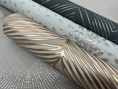 Three rolls of York Wallcoverings Polished Chevron Wallpaper, featuring metallic designs: one with gold and white abstract lines, one adorned with silver leaves on light gray, and one showcasing silver geometric lines on dark gray.