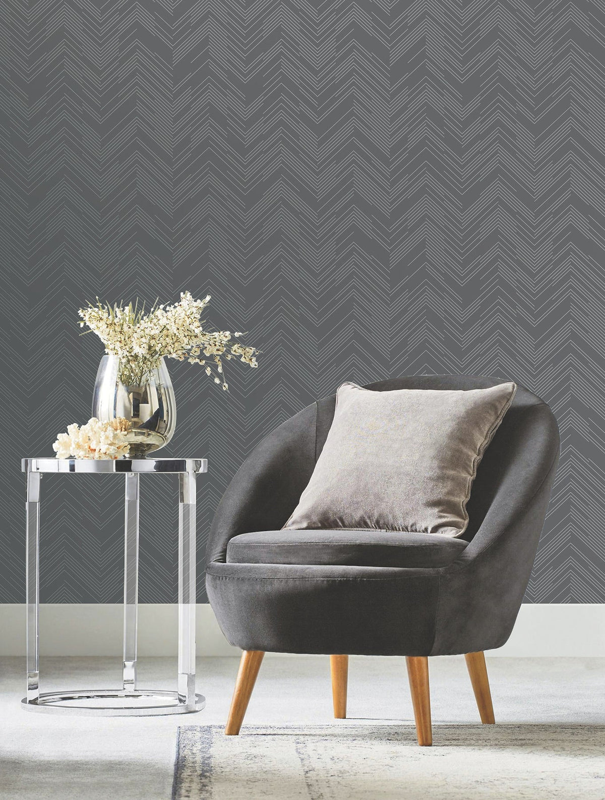 A gray modern armchair with a light cushion sits next to a round glass side table holding a vase of white flowers, set against Polished Chevron Wallpaper by York Wallcoverings.