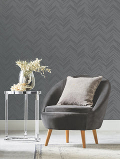 A gray modern armchair with a light cushion sits next to a round glass side table holding a vase of white flowers, set against Polished Chevron Wallpaper by York Wallcoverings.