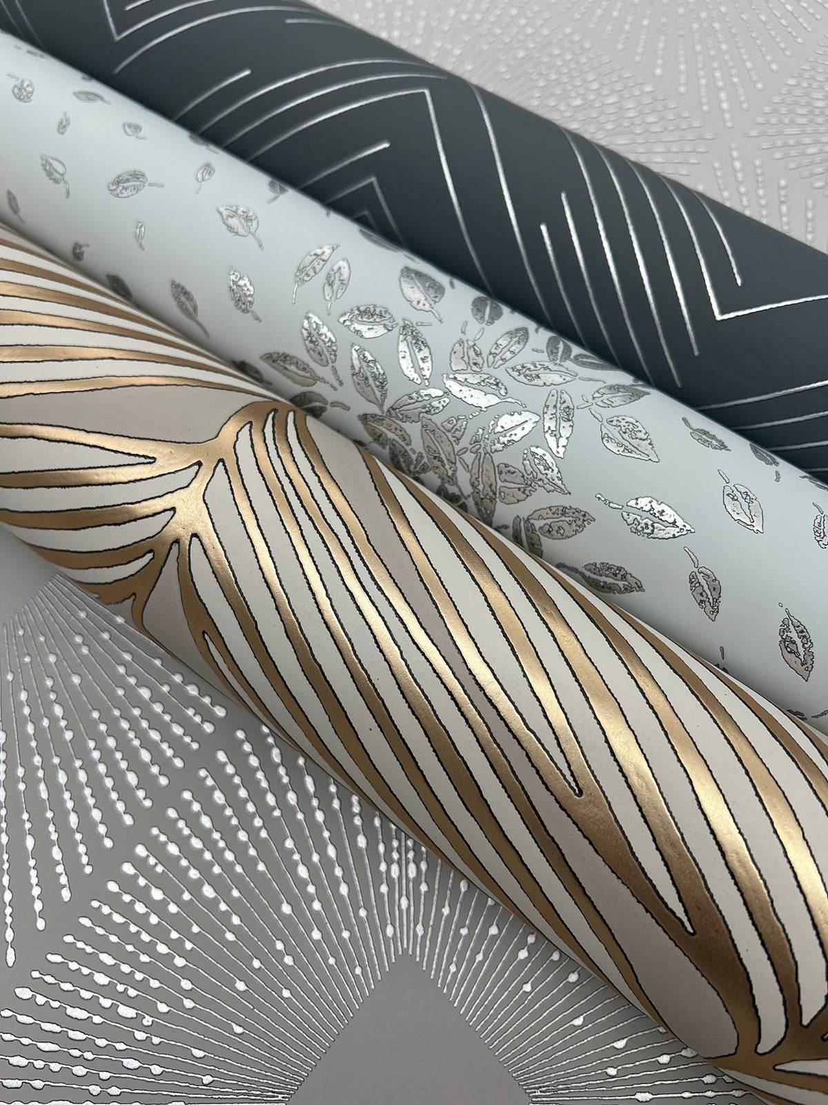 Three rolls of Polished Chevron Wallpaper by York Wallcoverings, each featuring metallic patterns: one dark roll with silver chevrons, one light roll with silver leaves, and one cream roll with gold and silver organic lines—all displayed on a grey patterned surface.