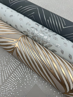 Three rolls of Polished Chevron Wallpaper by York Wallcoverings, each featuring metallic patterns: one dark roll with silver chevrons, one light roll with silver leaves, and one cream roll with gold and silver organic lines—all displayed on a grey patterned surface.