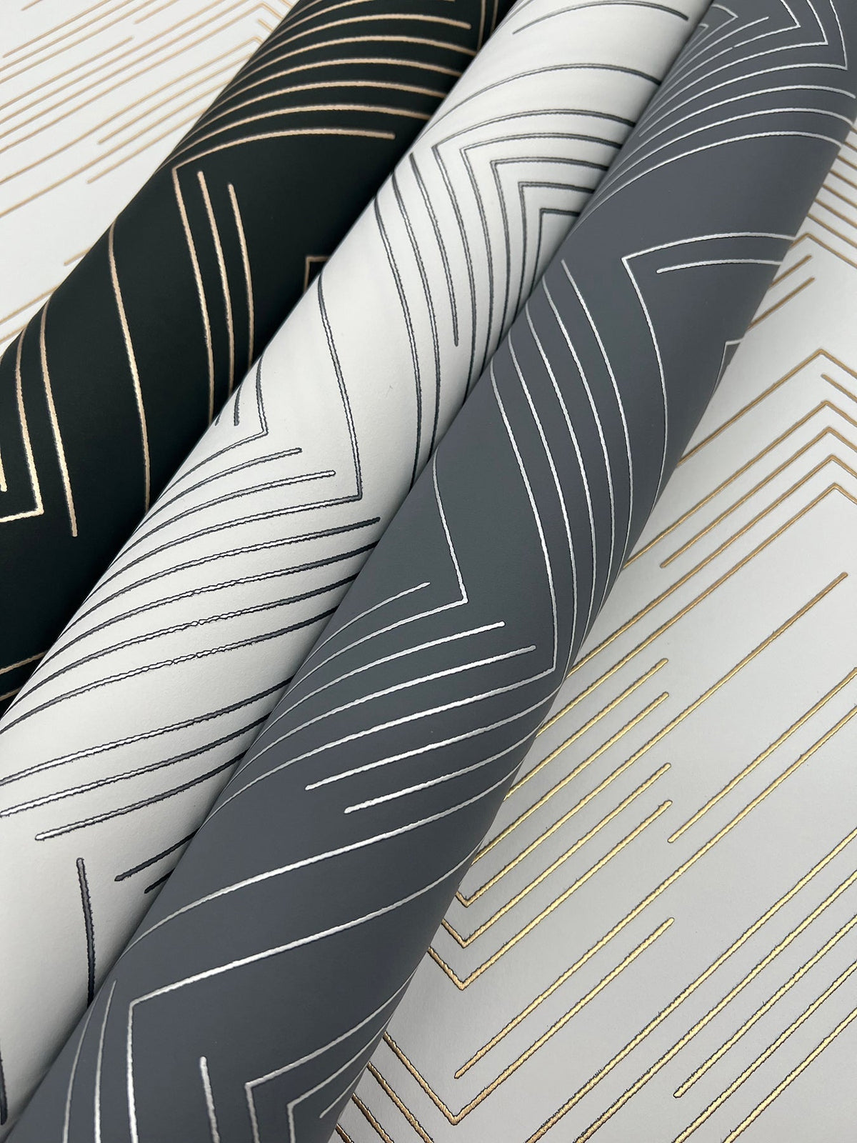 Three rolls of Polished Chevron Wallpaper by York Wallcoverings in black, white, and gray, each showcasing metallic geometric line patterns. The designs feature angular, intersecting chevron lines in gold or silver tones for a modern and elegant aesthetic.