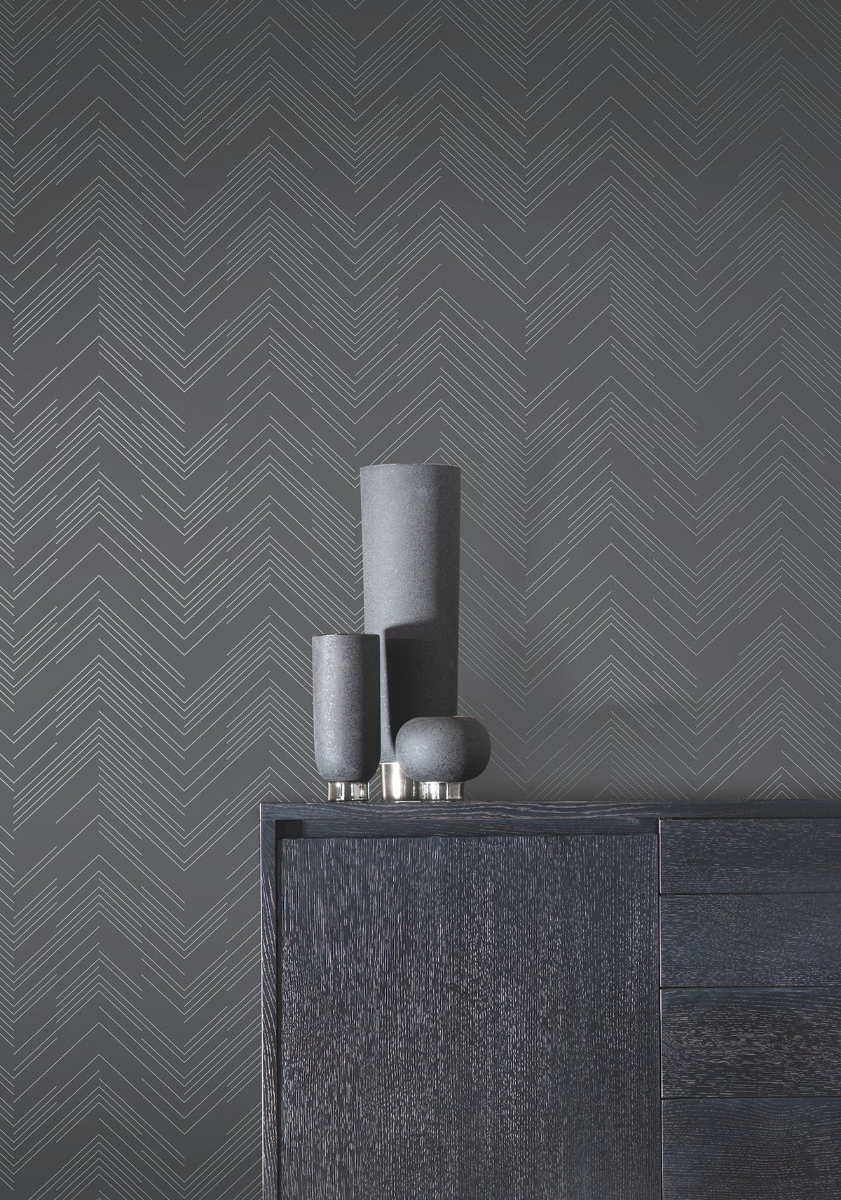 Three matte black vases sit on a dark wood cabinet, set against a gray wall adorned with York Wallcoverings’ Polished Chevron Wallpaper.
