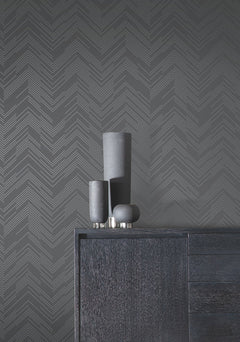 Three matte black vases sit on a dark wood cabinet, set against a gray wall adorned with York Wallcoverings’ Polished Chevron Wallpaper.