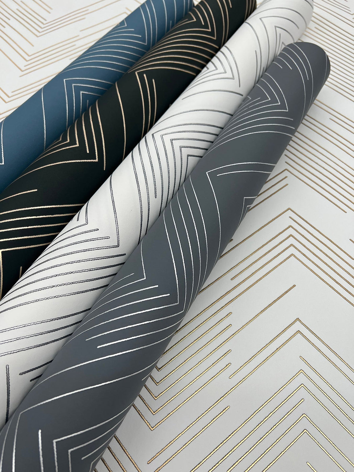 Four rolls of York Wallcoverings’ Polished Chevron Wallpaper, featuring metallic gold geometric line designs in blue, black, white, and gray colorways, are displayed on a flat surface adorned with a matching gold pattern.