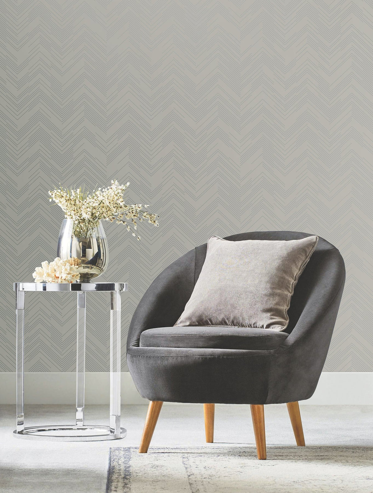 A modern gray armchair with a cushioned pillow sits beside a round glass side table holding a vase of white flowers, set against York Wallcoverings’ Polished Chevron Wallpaper and soft carpet.