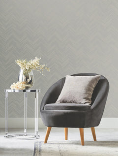 A modern gray armchair with a cushioned pillow sits beside a round glass side table holding a vase of white flowers, set against York Wallcoverings’ Polished Chevron Wallpaper and soft carpet.