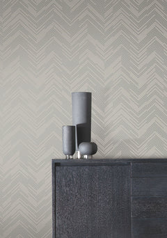 A modern black cabinet with three tall, dark vases and a small decorative item on top is set against a beige wall adorned with York Wallcoverings’ Polished Chevron Wallpaper in a subtle gray pattern.