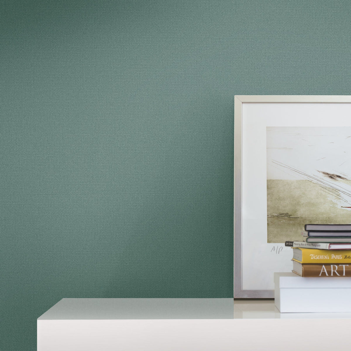 A minimalist and modern space features a green Chevronette Wallpaper by York Wallcoverings behind a white shelf displaying a framed artwork and a small stack of books, one labeled ART.