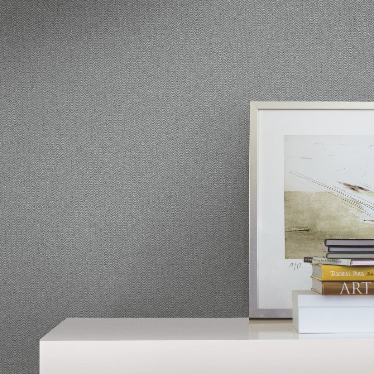 Framed artwork and stacked books sit on a white surface against a wall adorned with Chevronette Wallpaper by York Wallcoverings, creating a minimalist and modern decor scene.