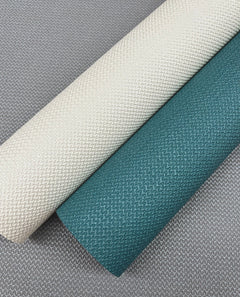 Two rolls of York Wallcoverings’ Chevronette Wallpaper, one in cream and the other in teal, lie side by side on a gray, woven surface featuring a similar textured pattern.