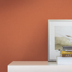 A framed abstract art print, stacked books, and a white box sit on a glossy white surface against a textured burnt orange wall covered in York Wallcoverings’ Chevronette Wallpaper.