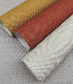 Three rolls of Chevronette Wallpaper by York Wallcoverings—one in gold, one in red, and one in silver—are arranged diagonally on a gray textured surface, displaying their subtle woven patterns.