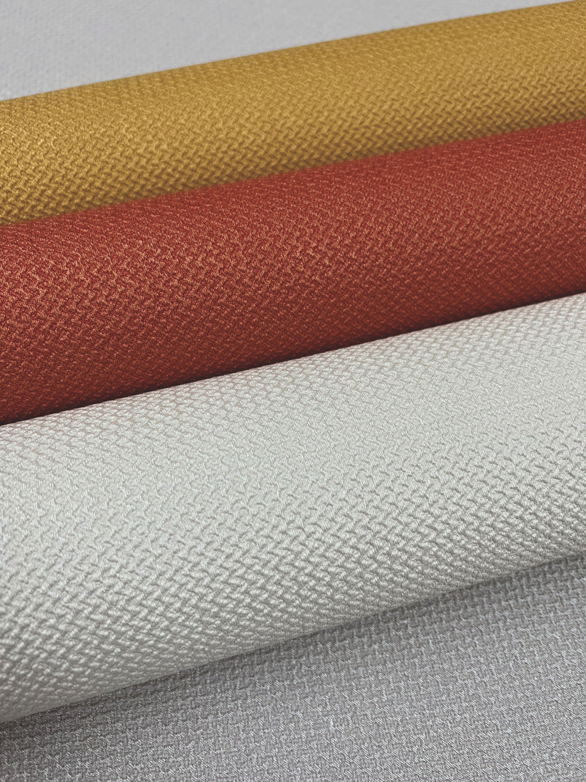 Four rolls of York Wallcoverings’ Chevronette Wallpaper in mustard yellow, burnt orange, and two shades of light gray are stacked diagonally, displaying their woven-inspired patterns.