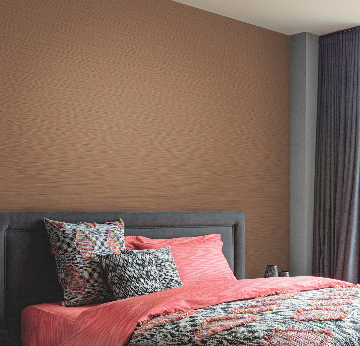 A modern bedroom featuring a padded dark gray headboard, coral pink bedding with abstract patterns, patterned pillows, and the Cannete Wallpaper by York Wallcoverings in light brown, complemented by dark floor-length curtains.