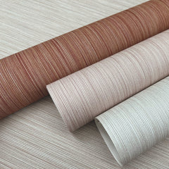 Three rolls of Cannete Wallpaper by York Wallcoverings in brown, beige, and off-white, displayed on a coordinating striped surface. The wallpapers feature subtle horizontal lines that evoke the look of woven fabric.