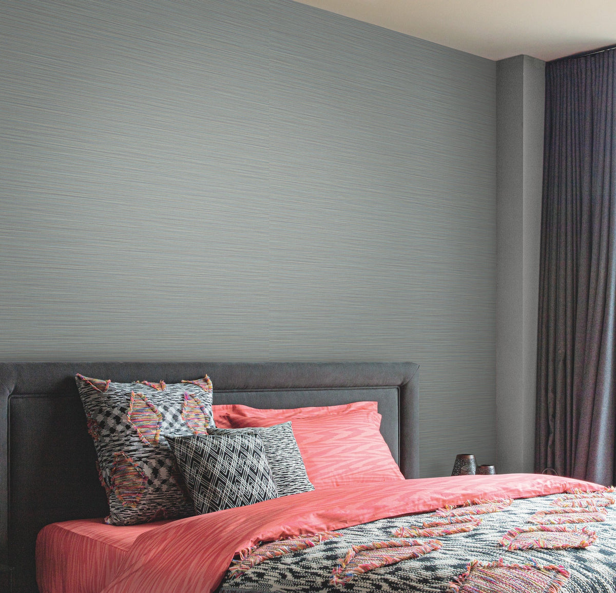 A bed with a dark upholstered headboard, coral and patterned bedding, and several pillows sits against a light gray York Wallcoverings Cannete Wallpaper, near a window with dark curtains.