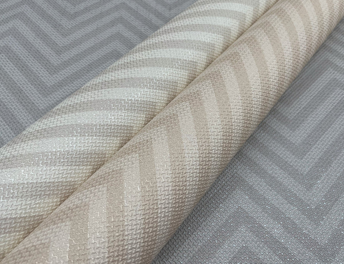 Two rolls of textured fabric with diagonal striped patterns, one in cream and the other in beige, are placed on York Wallcoverings’ Macro Chevron Wallpaper in a grey colorway with a chevron zigzag design.