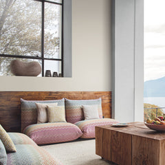 Modern living room featuring York Wallcoverings’ Macro Chevron Wallpaper on the neutral walls, low colorful cushions, a wooden coffee table, decorative vases, and large windows overlooking trees and a lake, creating a cozy and natural atmosphere.