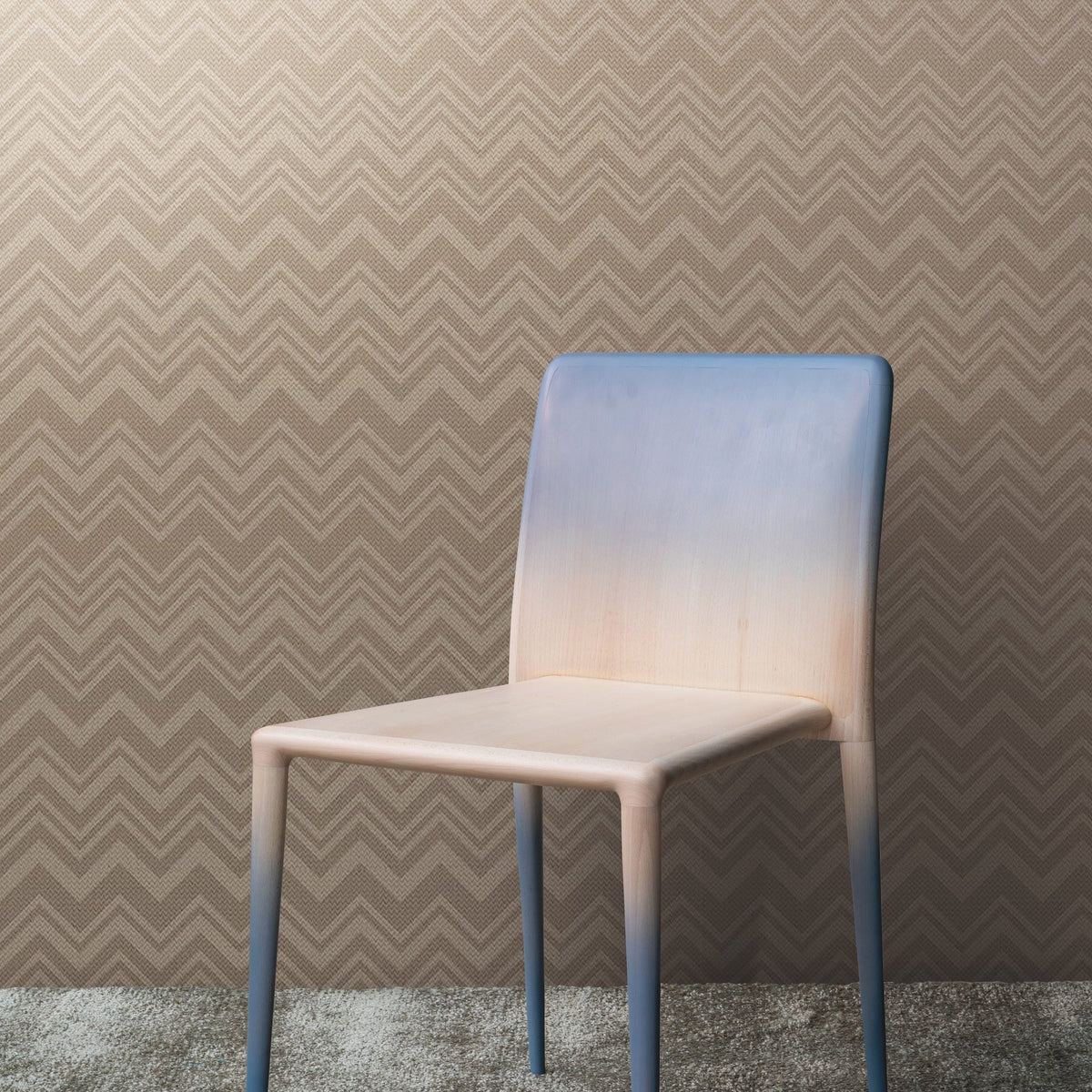 A modern chair with a gradient finish from light beige to blue sits on a textured carpet, in front of a wall adorned with Iconic Shades Wallpaper by York Wallcoverings, featuring a brown chevron pattern.